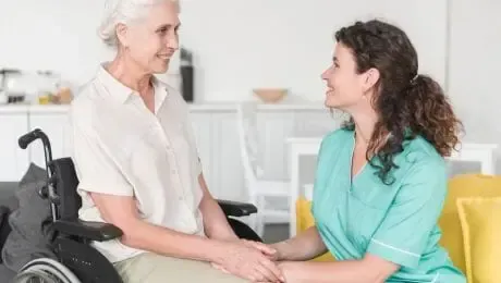 Woman in wheelchair and caregiver holding hands, smiling. Interior setting.