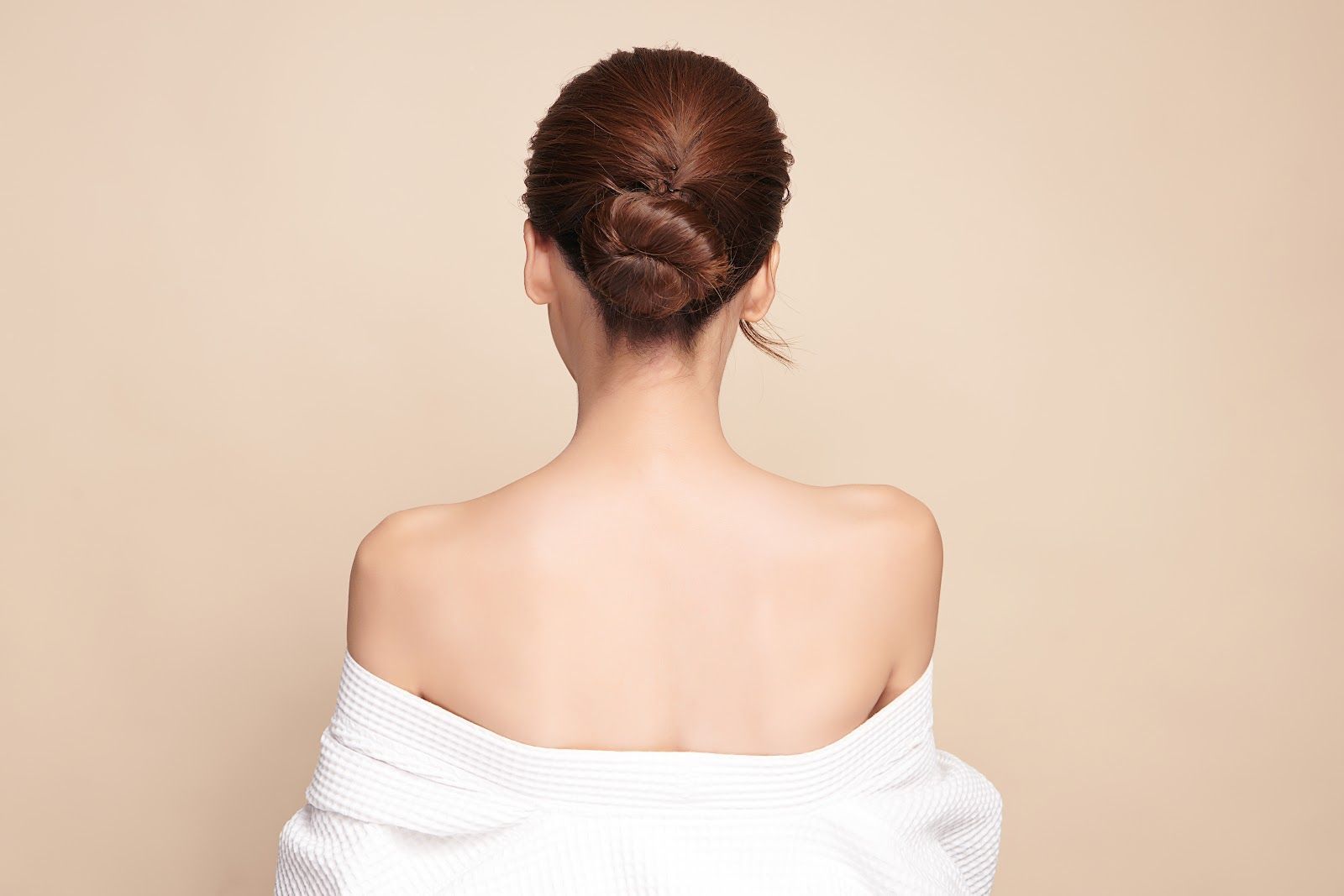 Back view of a person with hair in a bun, wearing an off-shoulder white top against a beige background