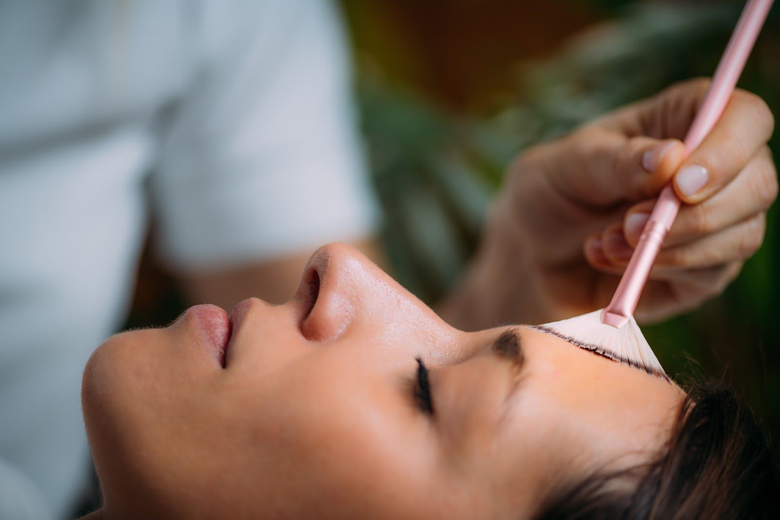 Person applies a pink facial mask with a brush to a woman lying down, eyes closed.