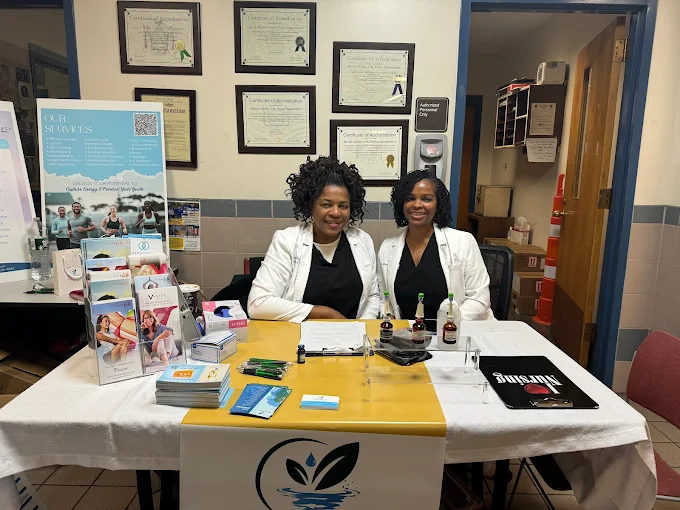 Two people in white coats sit at a table with informational brochures and a logo, in an office with wall-mounted certificates.