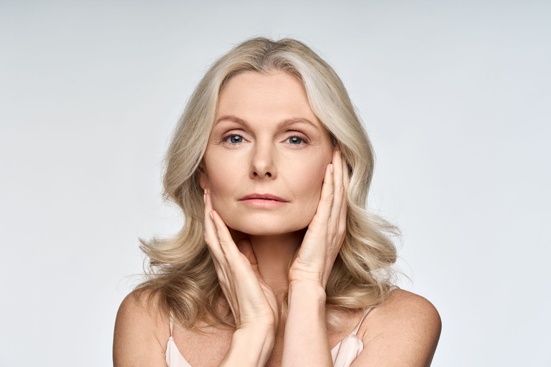 Woman with blonde hair touches her face with her hands, looking towards the camera.