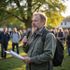 Man speaking into a microphone while holding papers at an outdoor gathering in a park