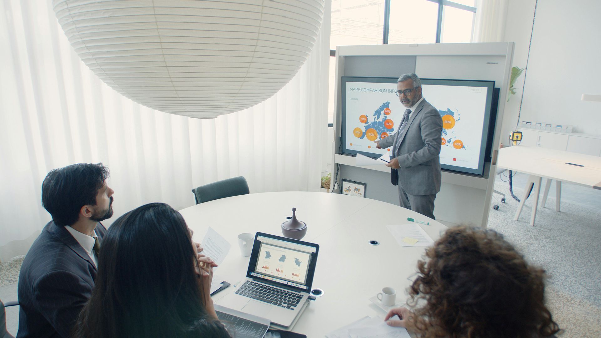 Meeting room presentation with a man presenting at a screen while three people watch around a table.