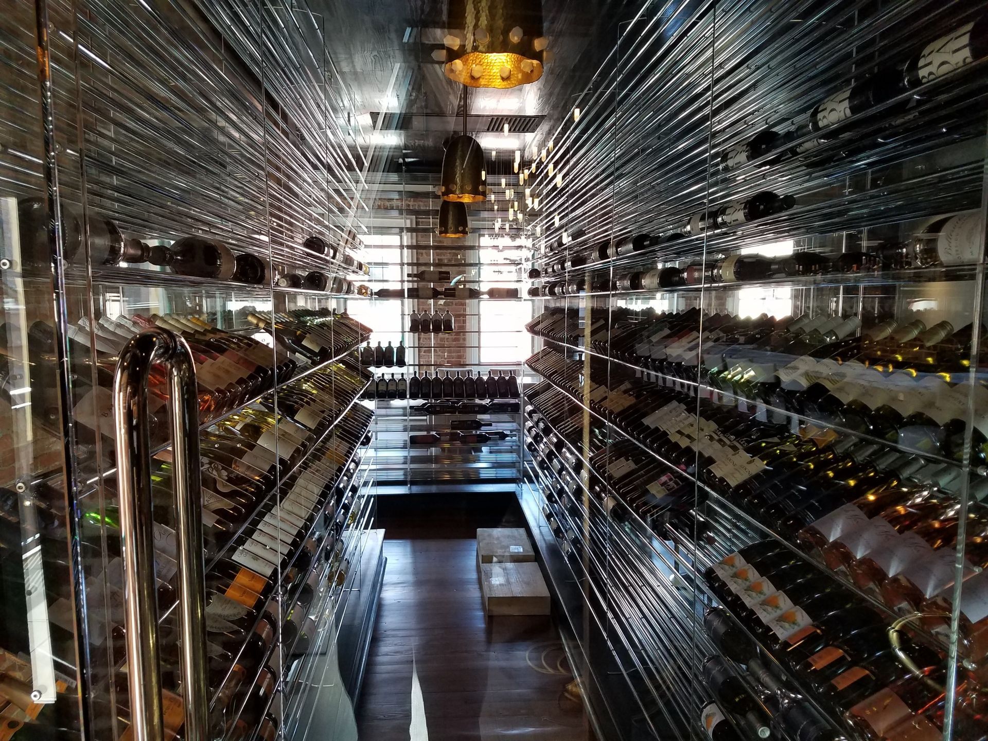 Glass-lined wine cellar with rows of bottles on both sides and a central aisle leading to a bright doorway