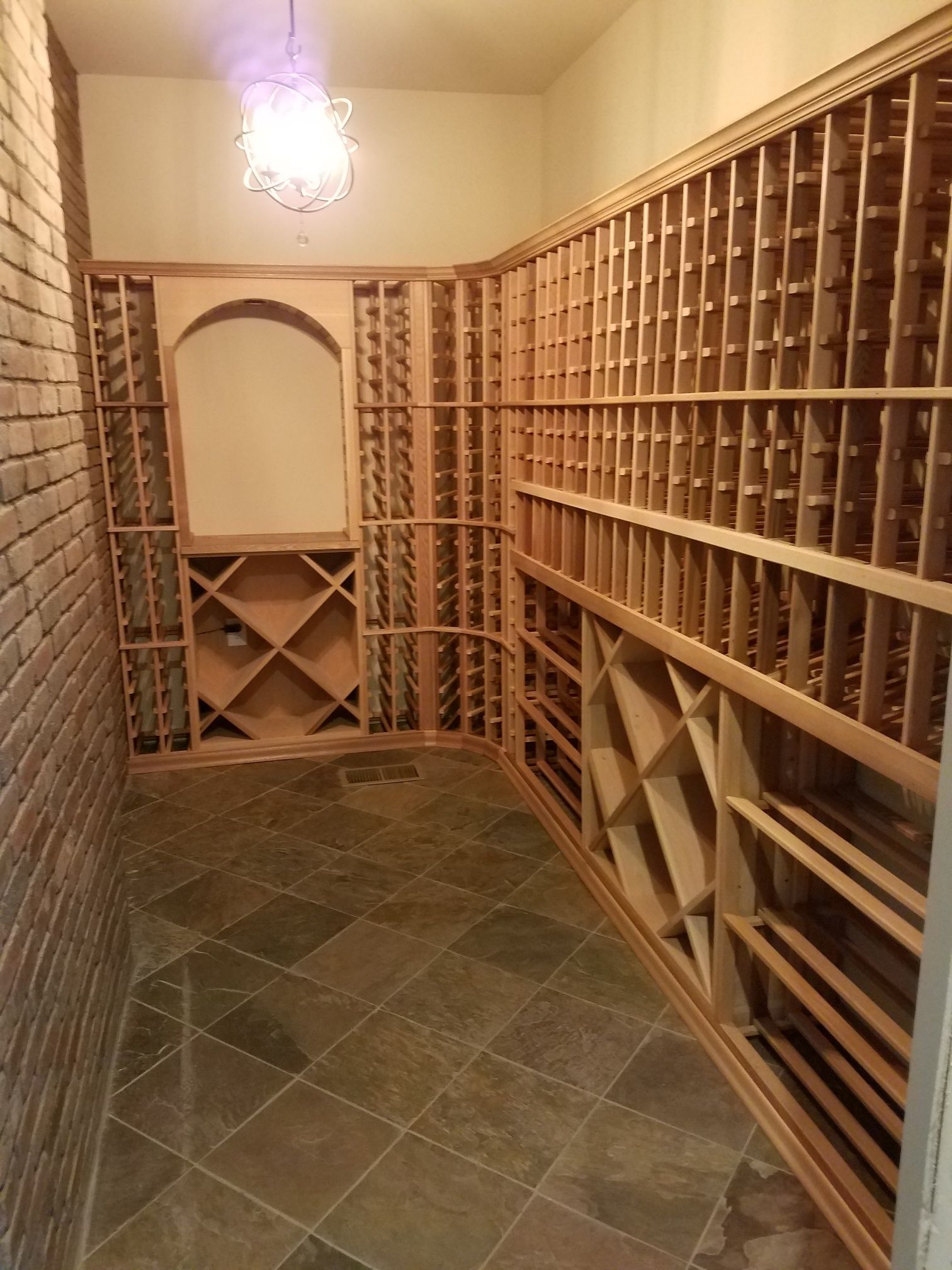 Wine cellar with wooden bottle racks lining both sides and a tiled floor under a hanging light.