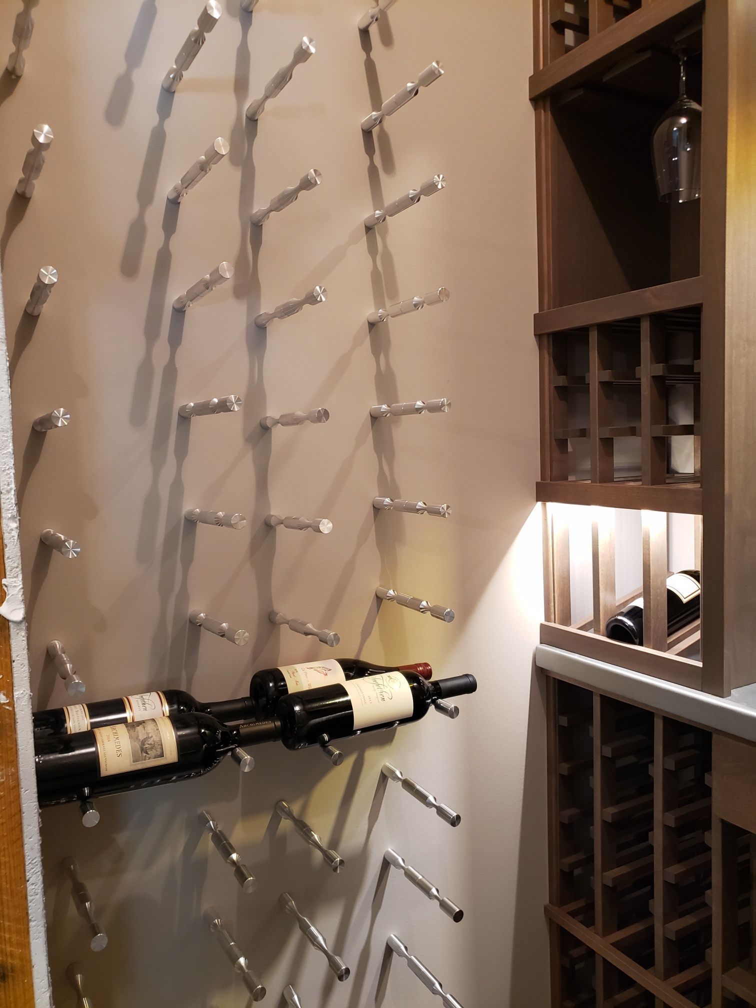 Wine bottles stored on a wall-mounted metal rack beside wooden shelving and a small lamp