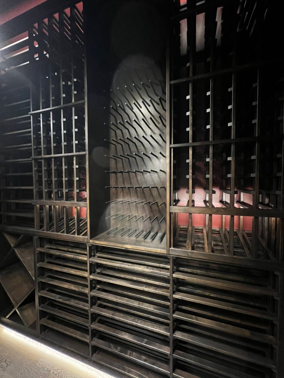 Dimly lit metal wine cellar with black racks, bottles, and a textured central column