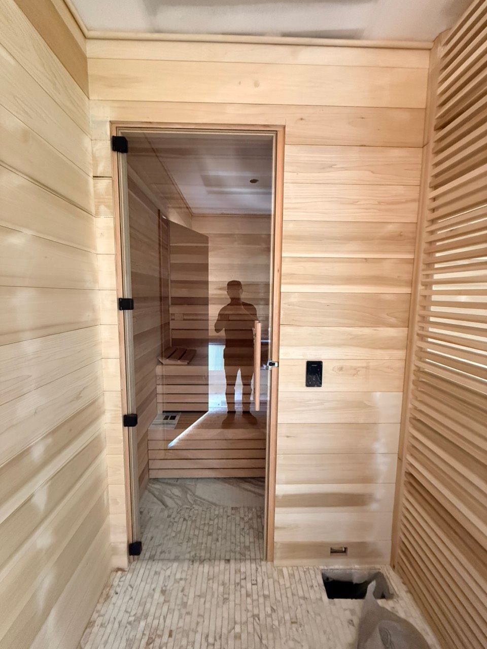 Wood-paneled sauna interior with glass door, heater vents, and a person reflected in the mirror.