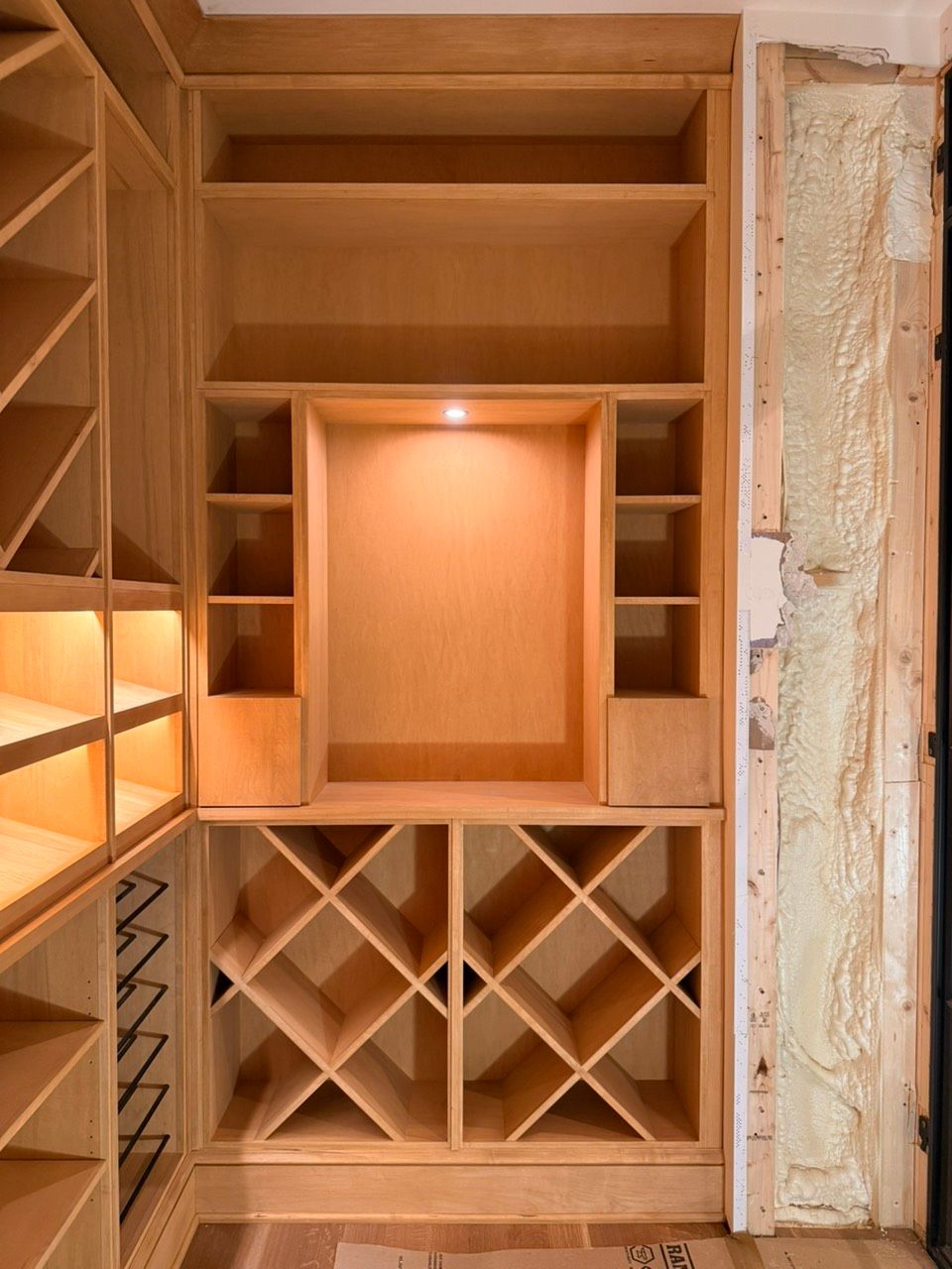 Built-in wooden shelving with wine racks and a lit display cubby in an unfinished wall niche