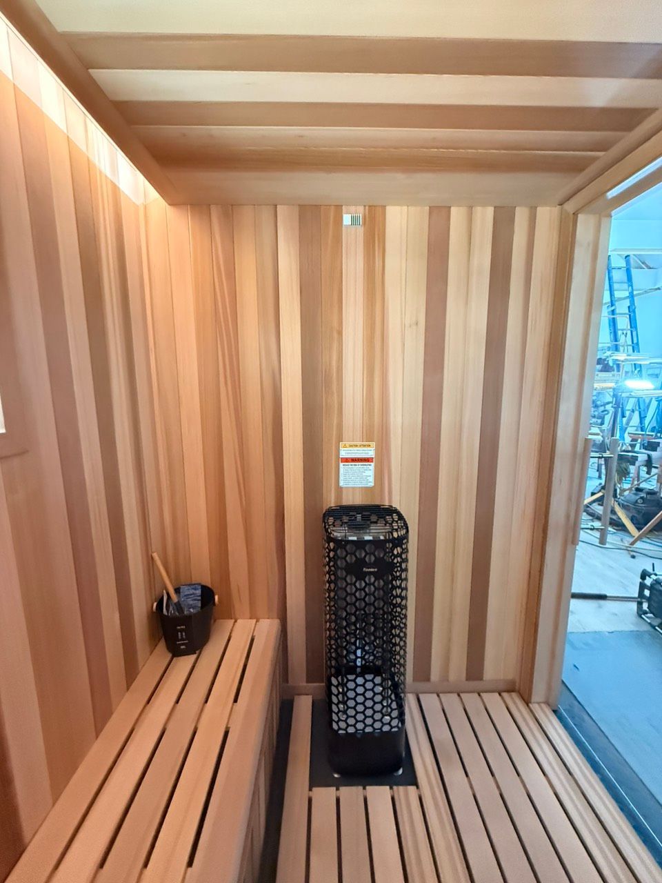 Wooden sauna interior with bench seating and a central heater, lit by daylight from the open doorway.