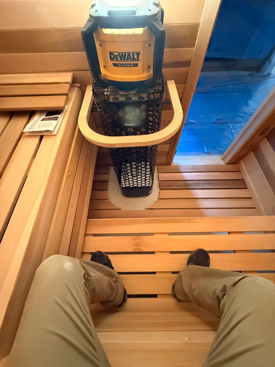 Inside a wooden sauna, a person sits on a bench facing a portable heater and a blue-lit window.