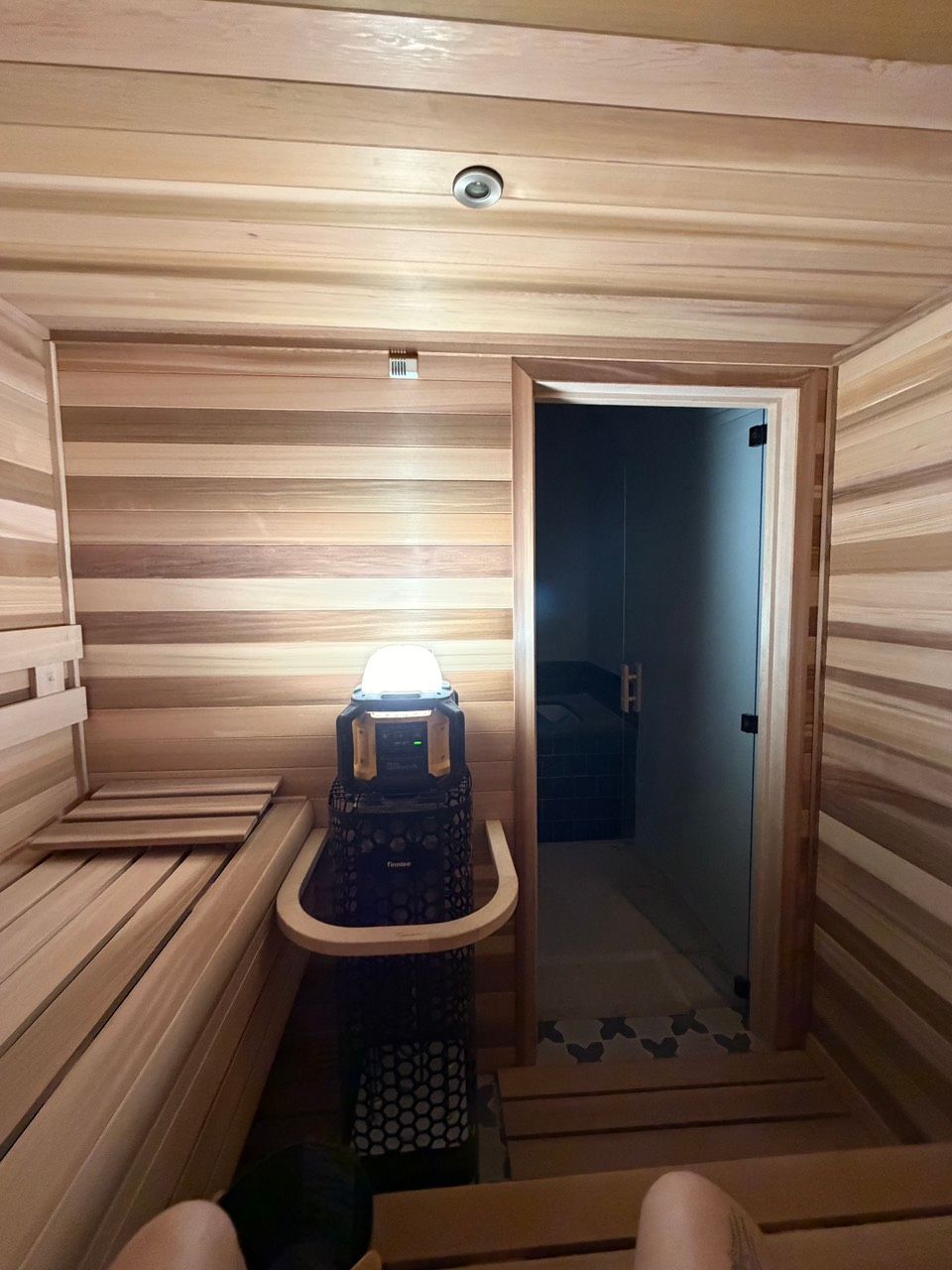 Wood-paneled sauna interior with bench seating, a heater, and an open doorway to a dim hall.
