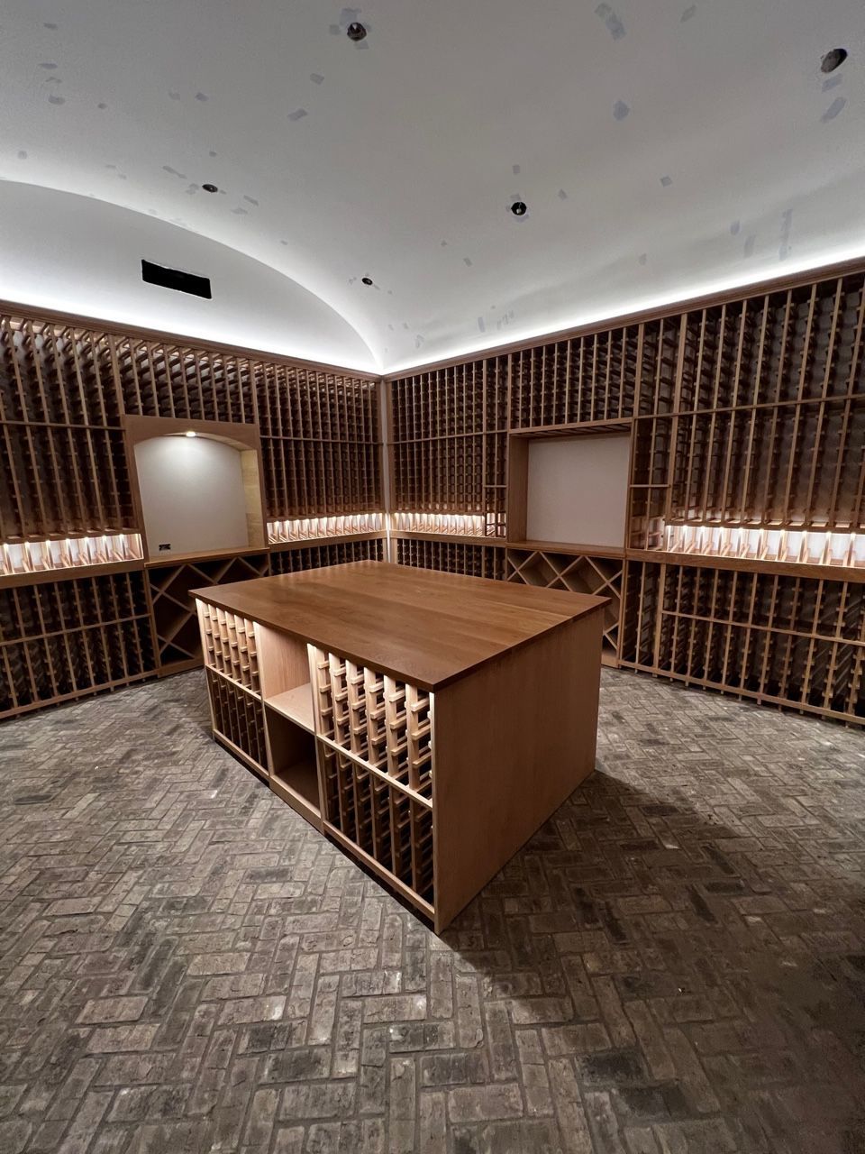 Empty wine cellar with wooden tasting table, floor-to-ceiling bottle racks, and soft ceiling lighting