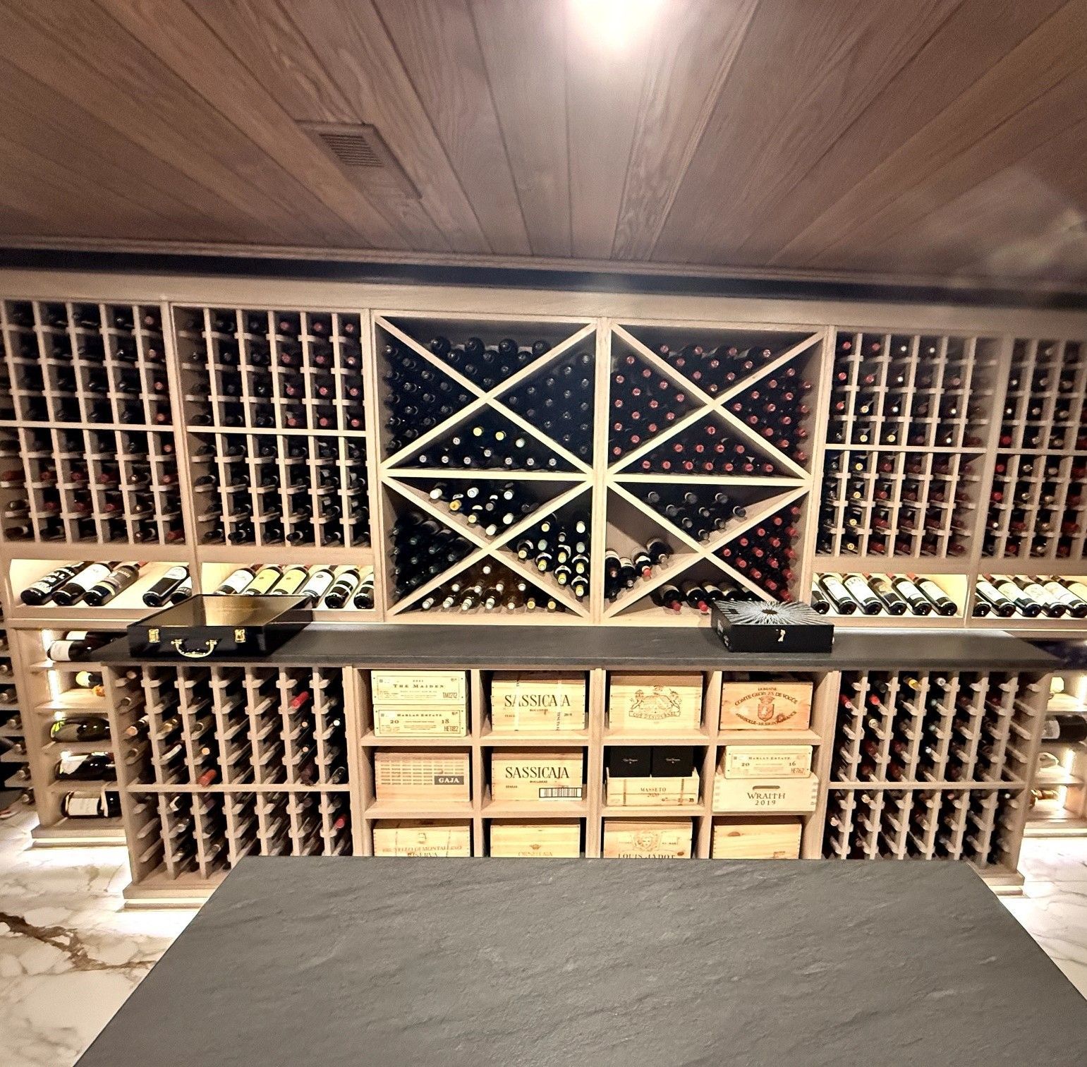 Wall-mounted wine rack with bottles, labeled storage drawers, and a dark countertop in a wood-paneled room