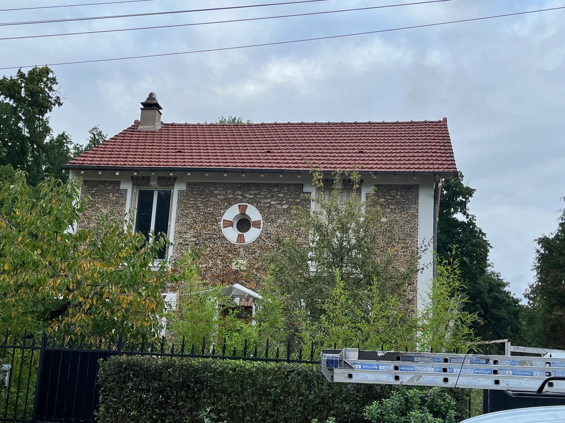 Maison en pierre au toit de tuiles rouges, entourée d'arbres verts sous un ciel couvert.