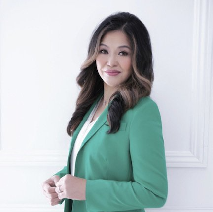 A woman in a green jacket is standing in front of a white wall.