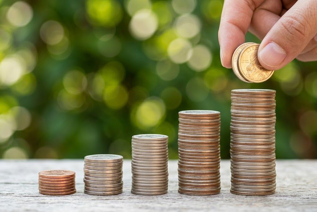 Hand placing a coin on a growing stack of coins on a wooden surface, with blurred green background