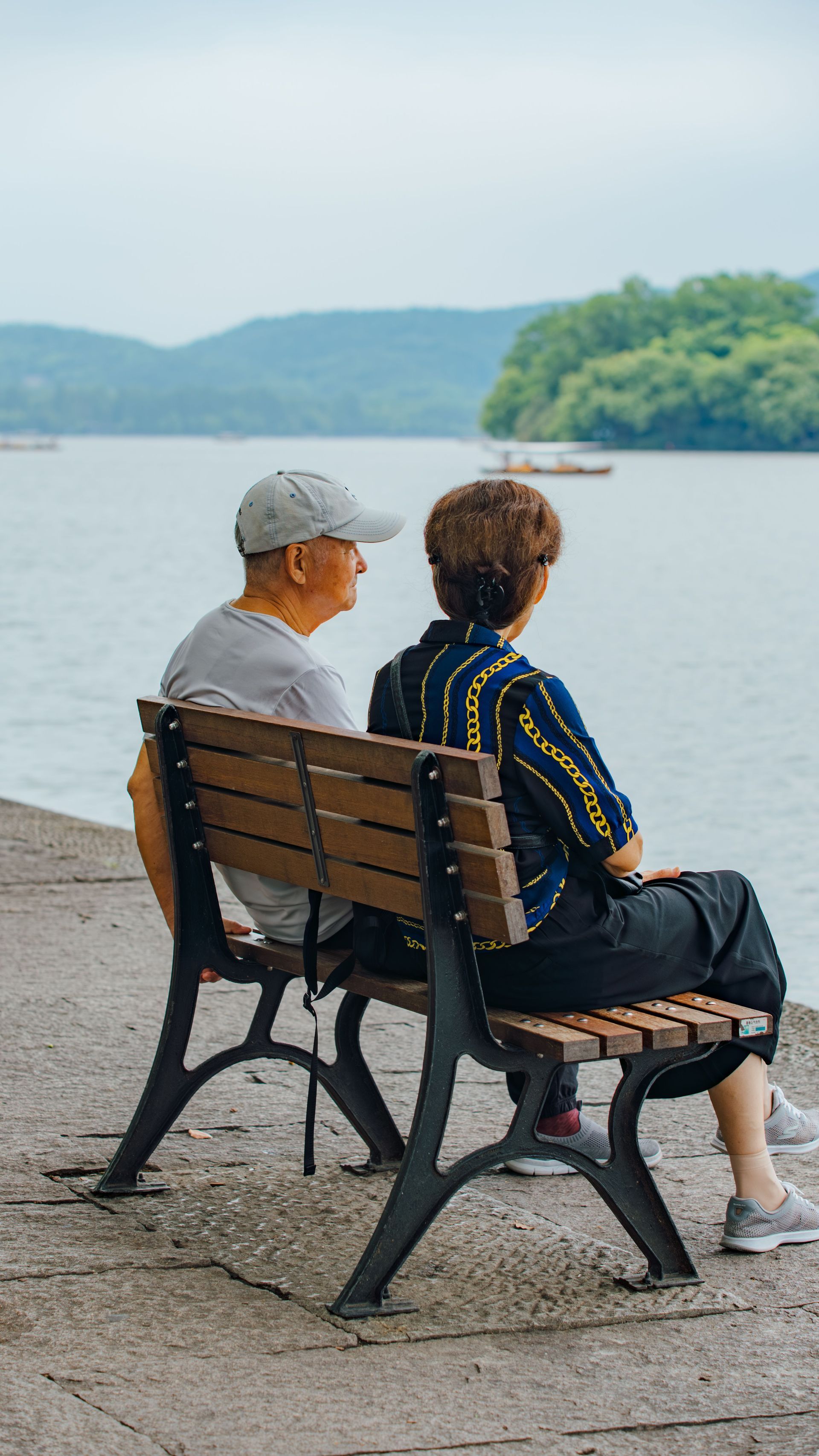 Two people sit on a bench by a calm lake, looking out at the water and distant hills.