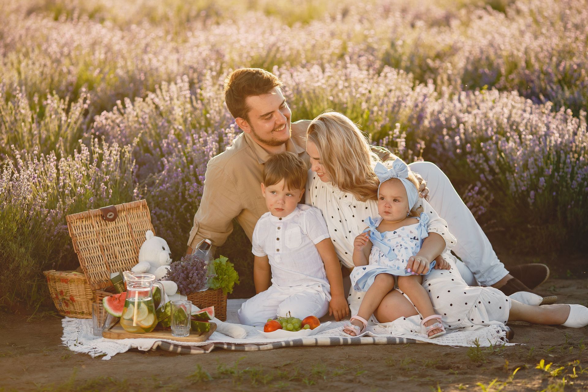 Family picnic in a lavender field, with a man and two children sitting on a blanket beside a basket and tea set.