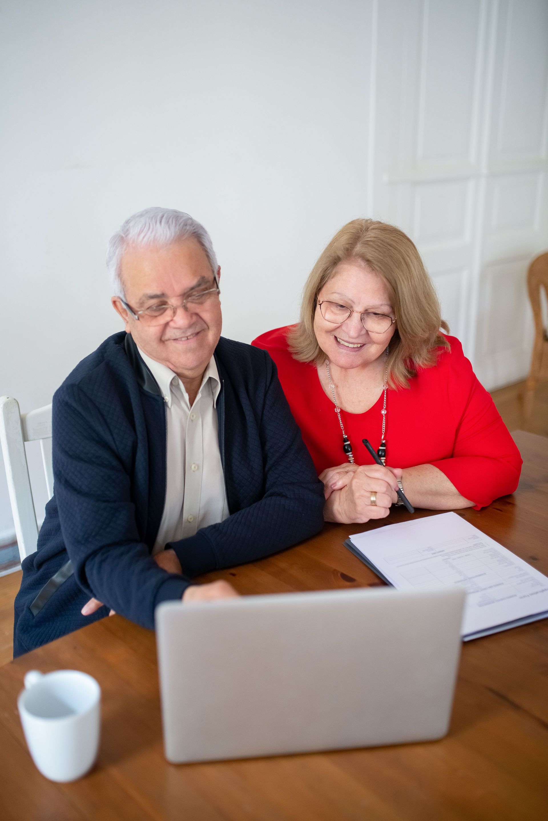 Two people smiling at a laptop on a wooden table with papers and a mug