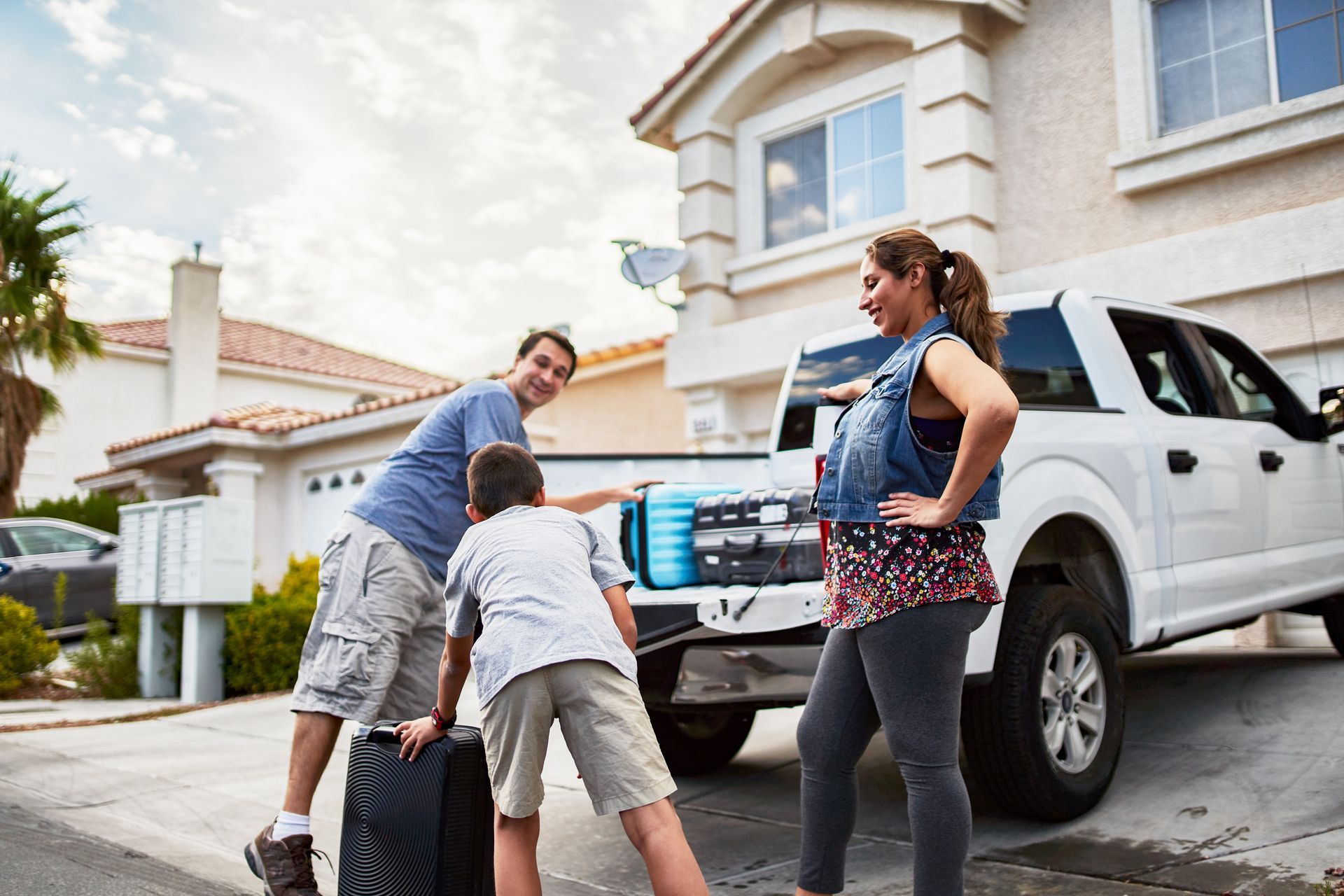 Two people unload luggage from a white pickup truck in a suburban driveway.