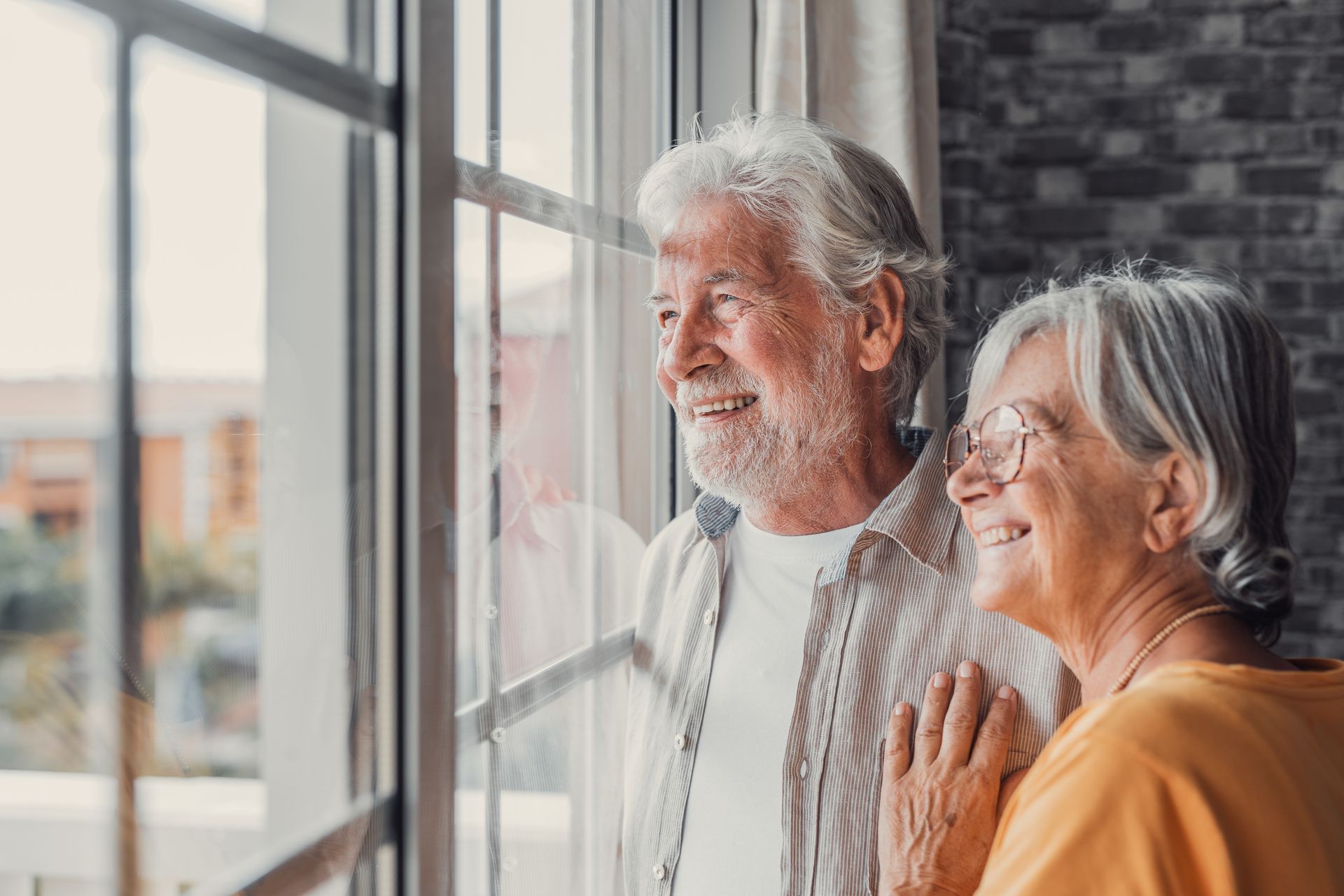 Two smiling people stand by a window, looking outside in warm light.