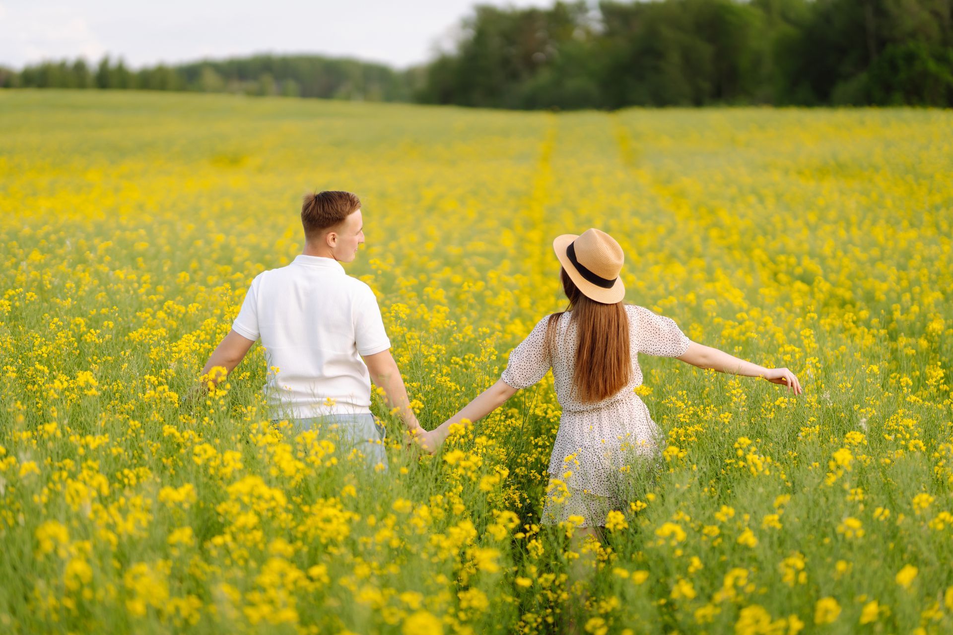 Couple holding hands in a yellow flower field, walking toward a green tree line