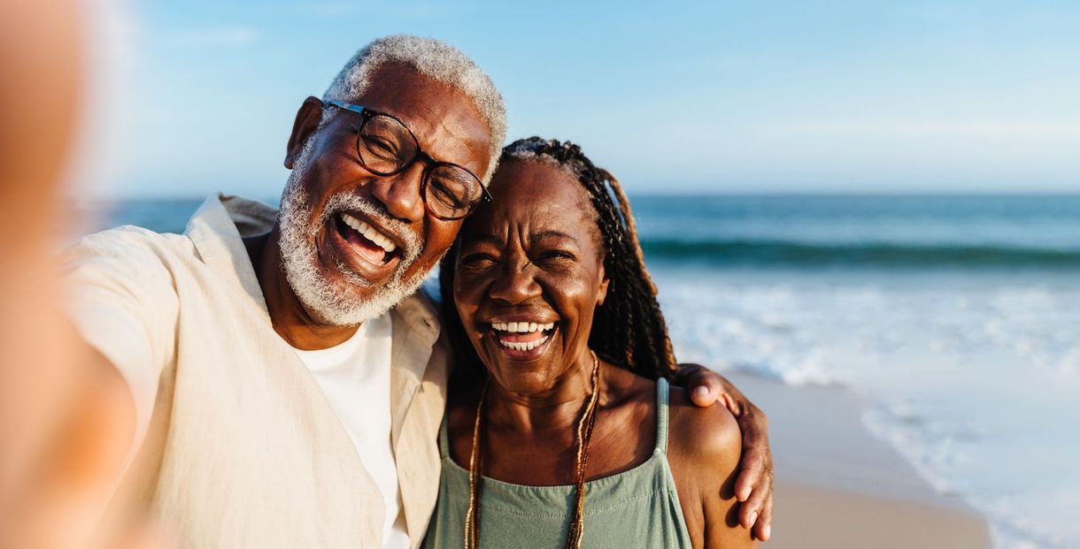 Two smiling people taking a selfie on a sunny beach with the ocean behind them