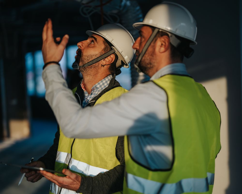 Two construction workers in hard hats and high-vis vests reviewing plans on a job site.