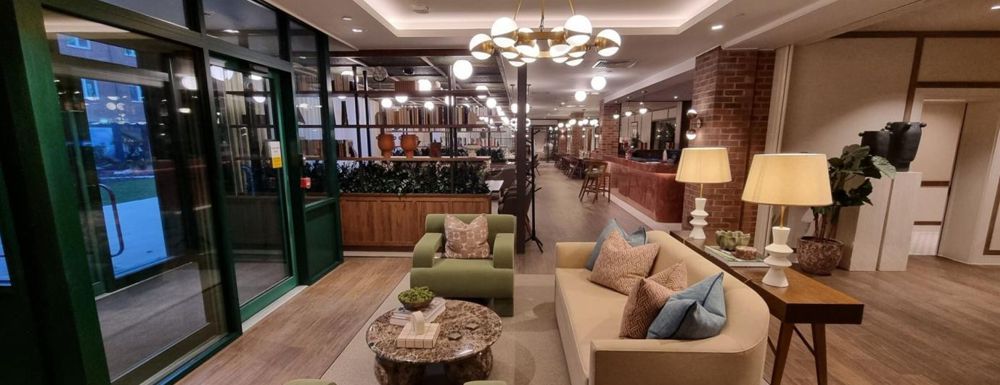 Modern lobby with beige sofas, green chairs, warm lighting, and floor-to-ceiling windows overlooking greenery