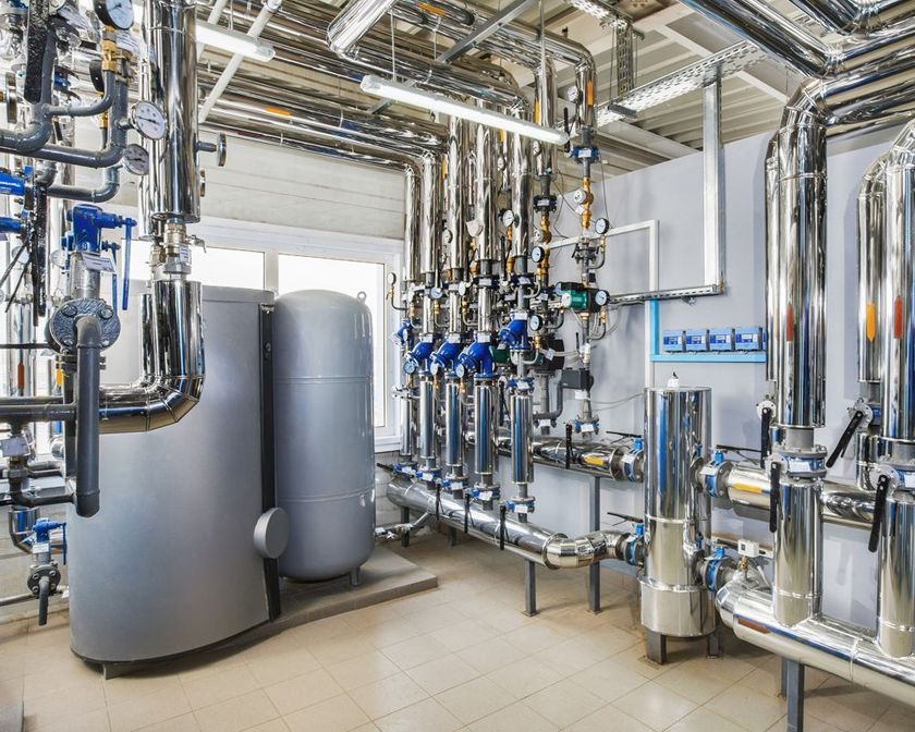 Industrial plant room with stainless steel pipes, valves, and large gray tanks inside a bright facility.