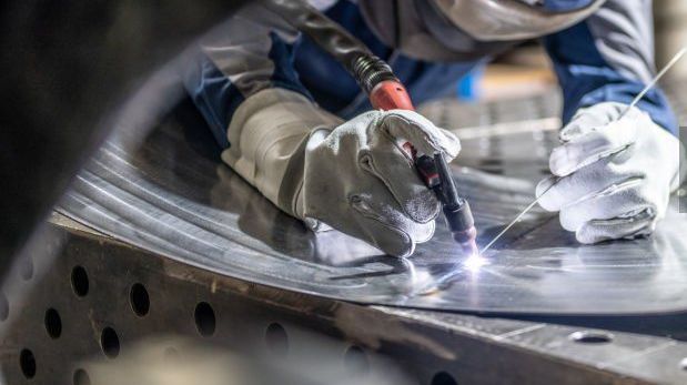 Welder in gloves welds metal with a bright arc in an industrial setting.