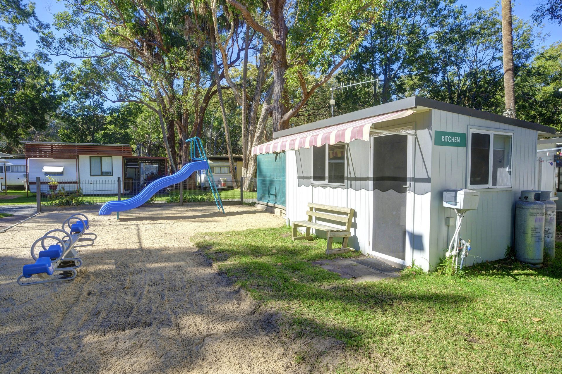 A playground with a slide and seesaw next to a small white cabin. Trees are in the background.— Pacific Palms Caravan Park in Elizabeth Beach, NSW