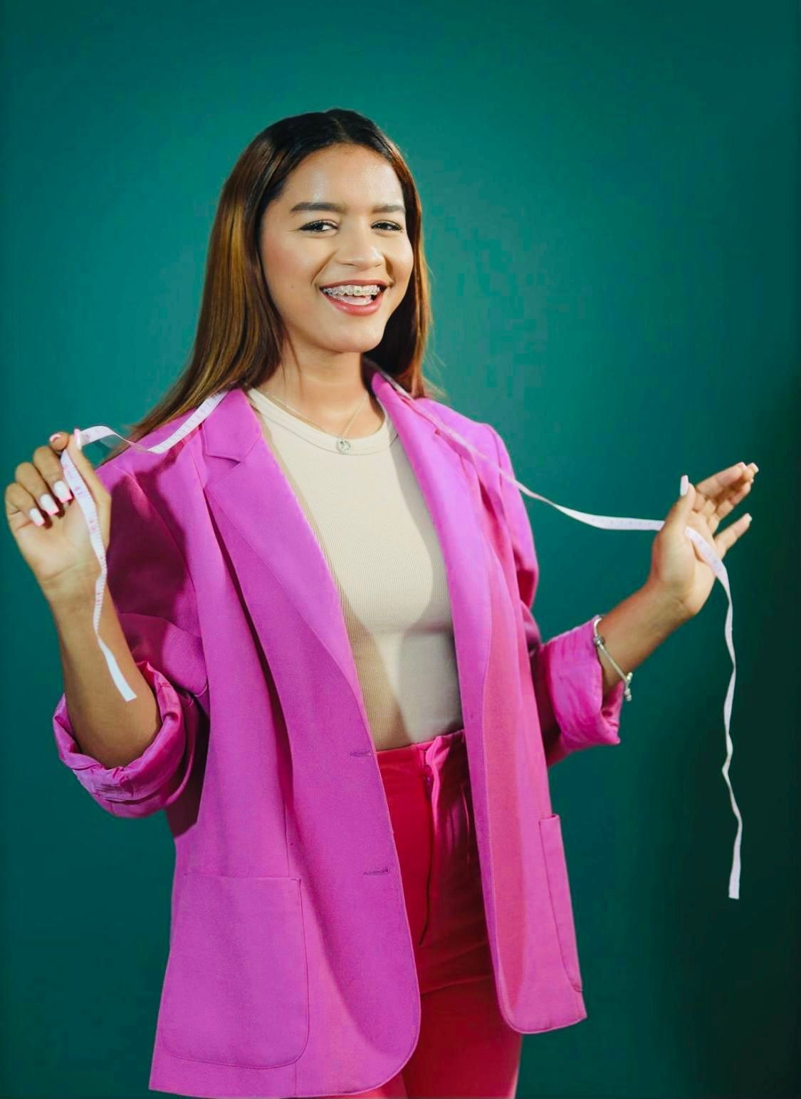 Smiling person in a pink blazer holds a white measuring tape against a teal background.