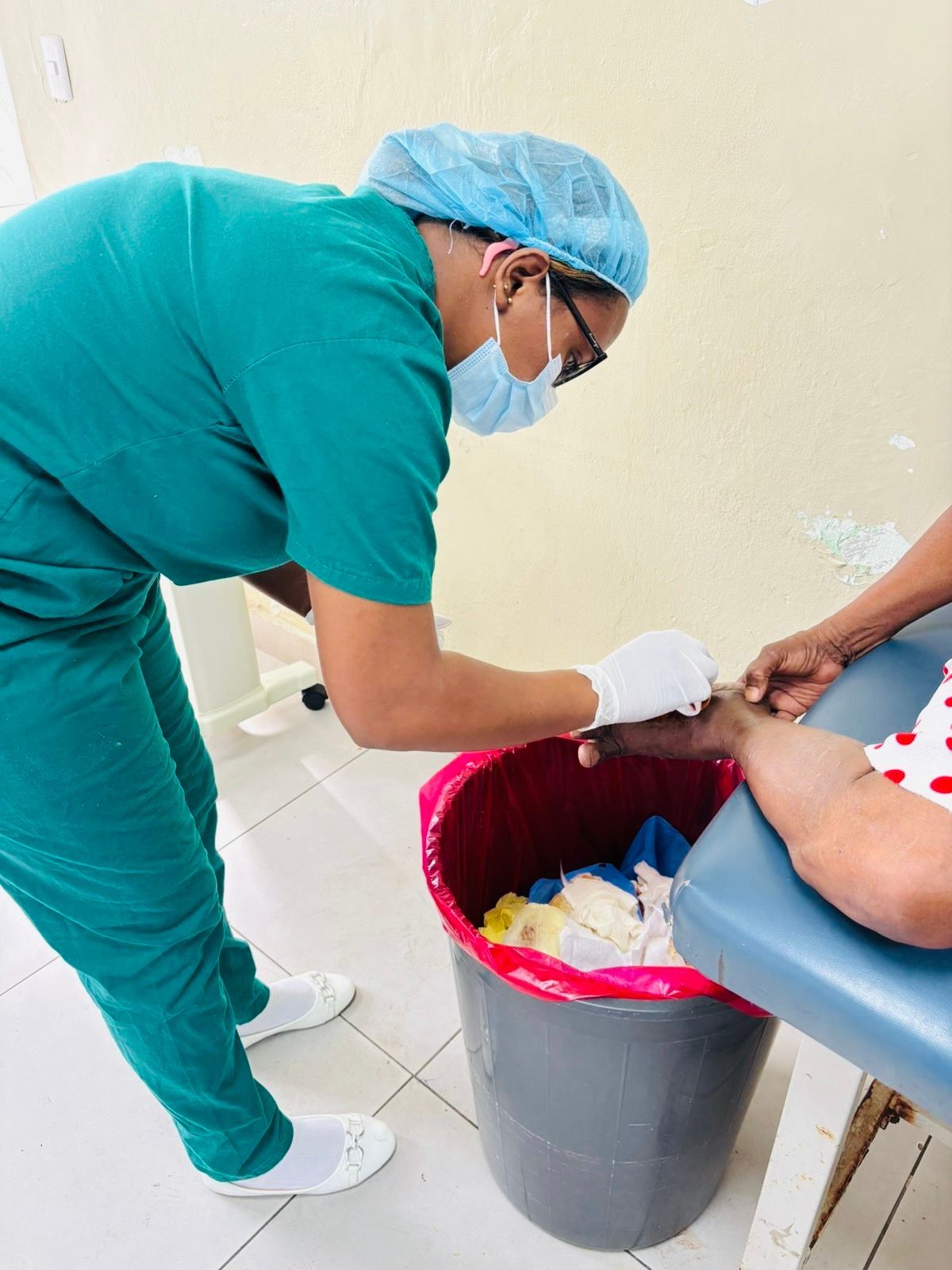Healthcare worker in teal scrubs tending to a patient's hand beside a red bin in a clinic room
