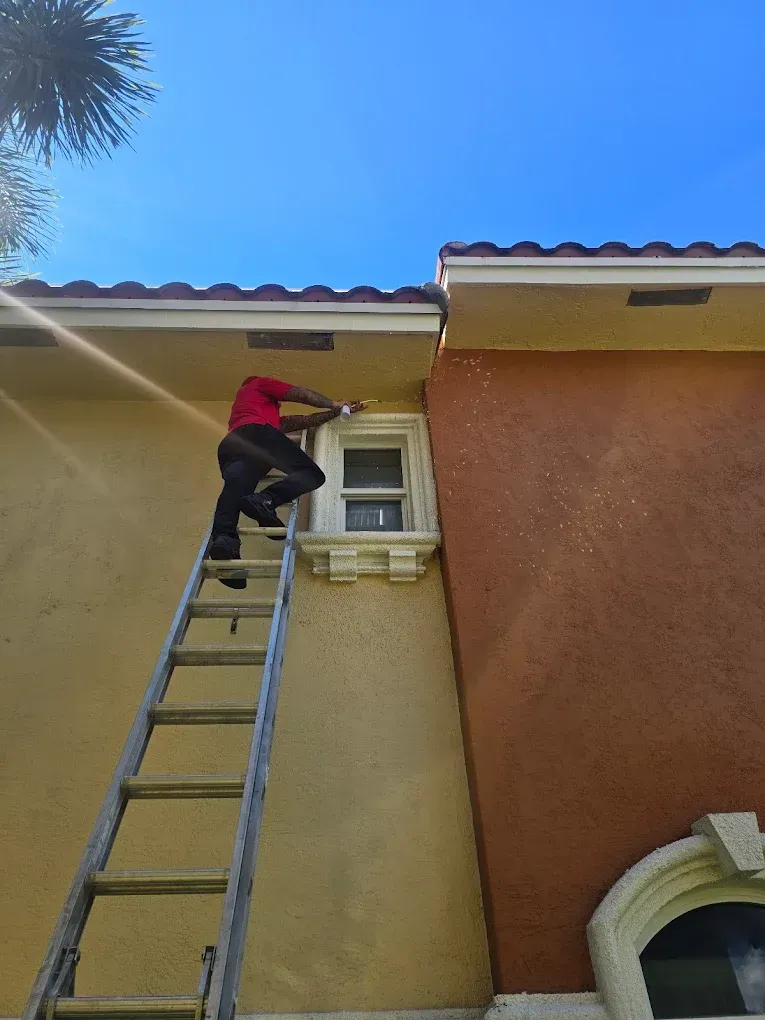 A person in a red shirt stands on an extension ladder, working on the exterior of a stucco home under a blue sky.