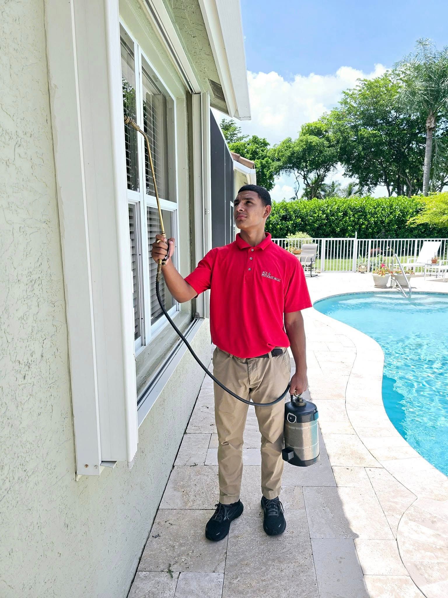 A person in a red uniform spraying a chemical treatment along the window frame of a home beside a swimming pool.