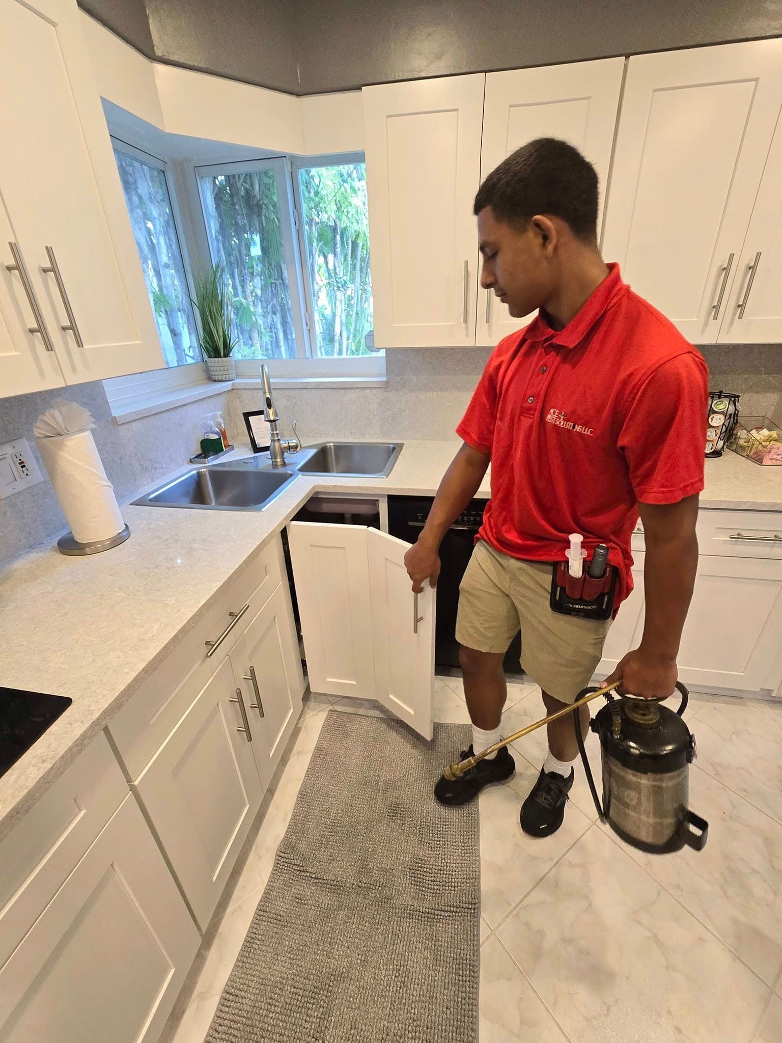 A pest control worker in a red shirt treats the kitchen under a sink with a handheld sprayer.