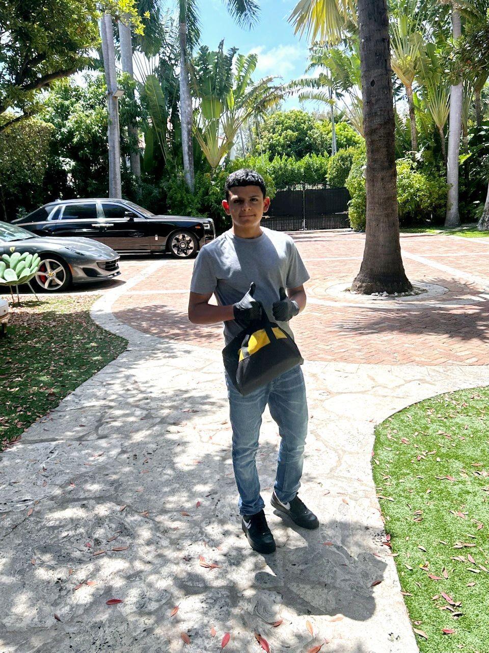A person in work clothes and gloves stands on a paved driveway holding a yellow and black tool bag near parked cars.