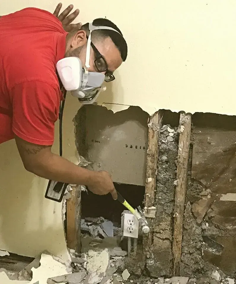 A person wearing a red shirt and a respirator mask using a small hammer to remove drywall near an electrical outlet.