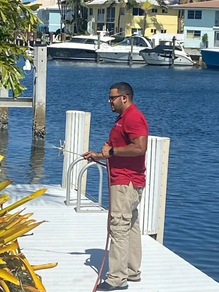 A person wearing a red polo shirt and tan cargo pants uses a pressure washer on a white boat dock by the water.