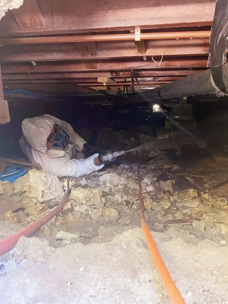 A worker in protective gear sprays foam insulation under the crawlspace of a wooden floor structure.