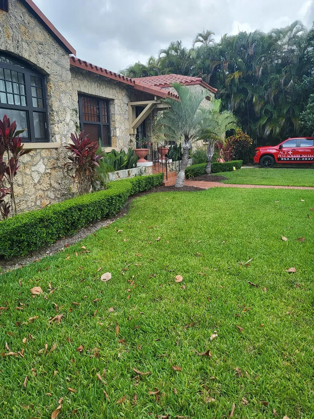 A stone-walled house with a red tile roof and a neatly trimmed hedge sits behind a green lawn, with a red truck parked.
