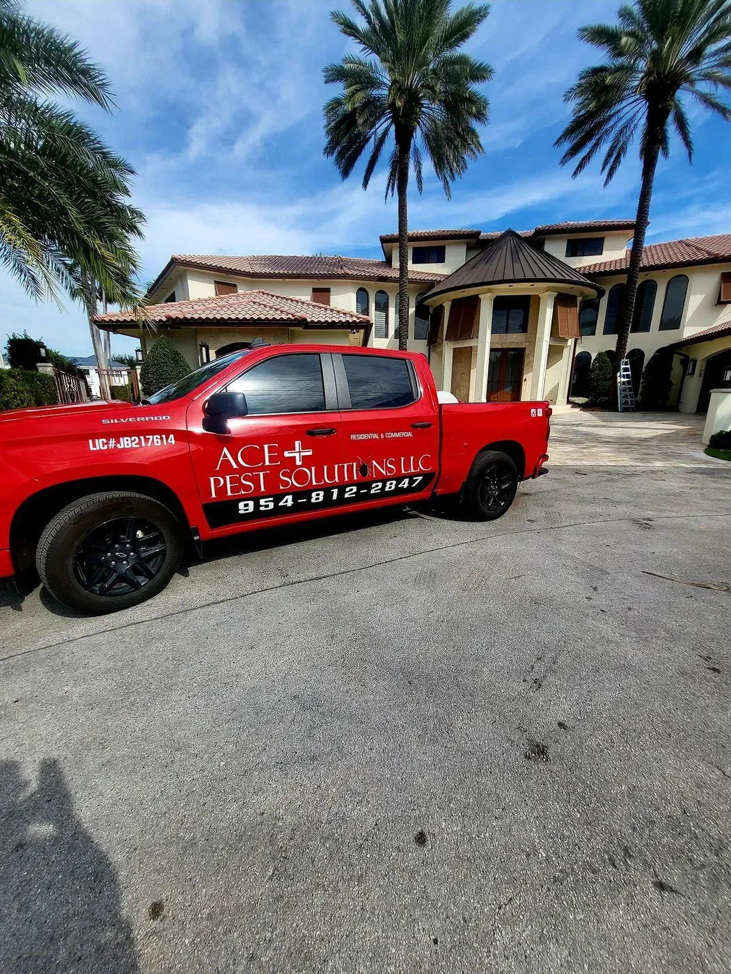 A red ACE Pest Solutions truck parked on a gravel driveway in front of a large, multi-story villa with palm trees.