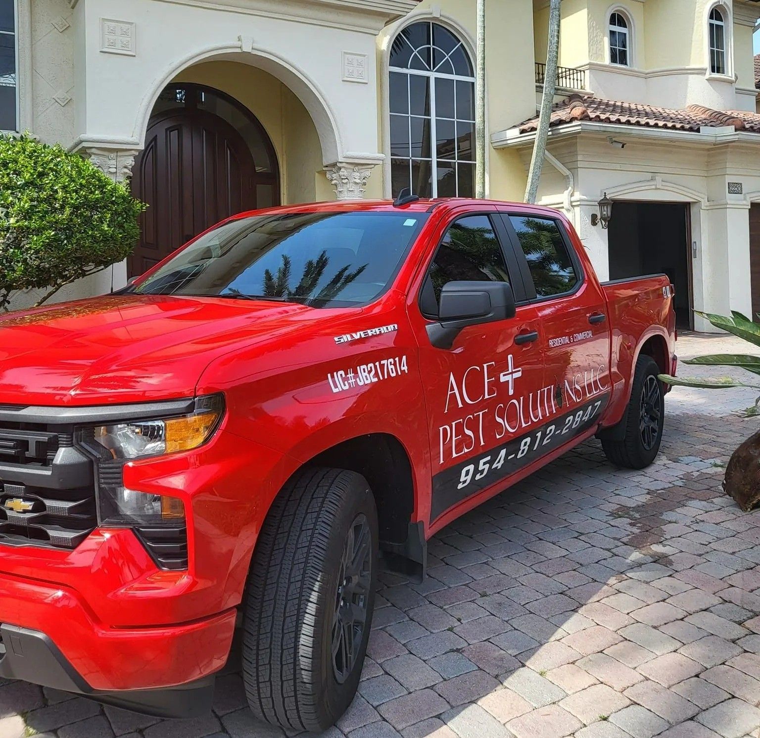 A bright red pickup truck for Ace Pest Squad parked in a stone-paved driveway in front of a tan residential home.