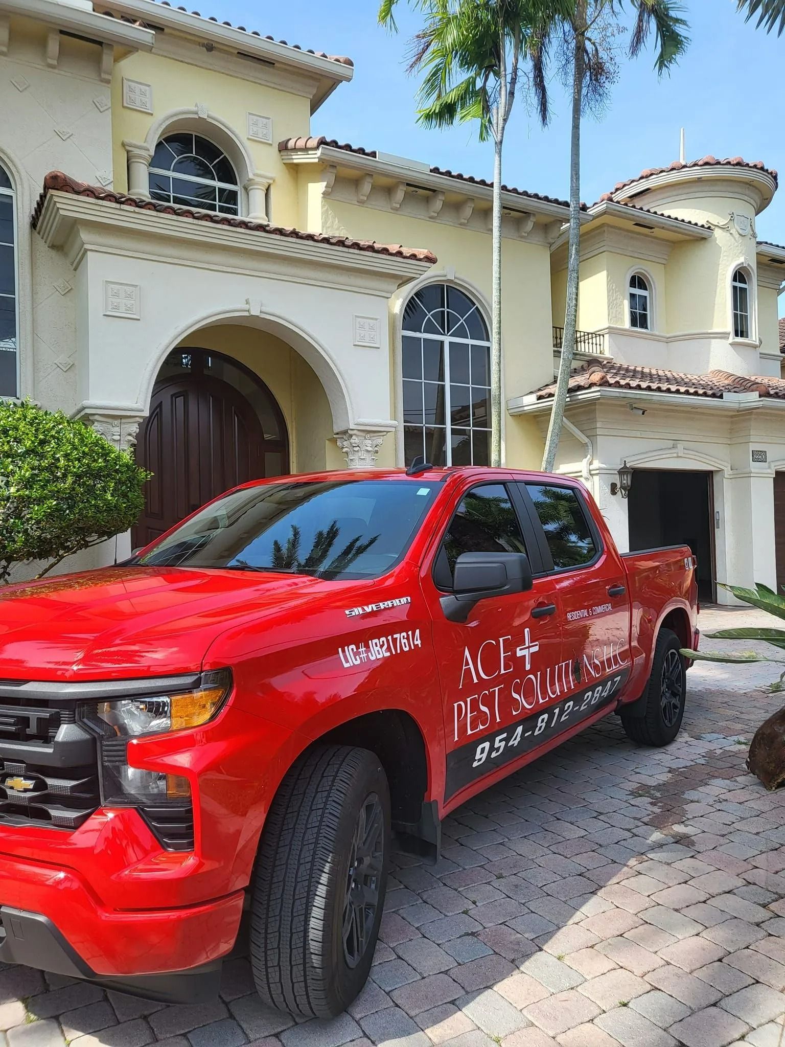 A bright red pickup truck parked in front of a large, light-yellow Mediterranean-style house on a sunny day.