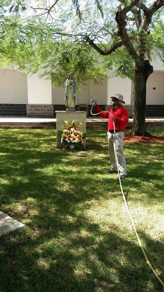 A person in a red shirt and hat sprays water from a hose at a statue of a figure on a pedestal in a grassy yard.