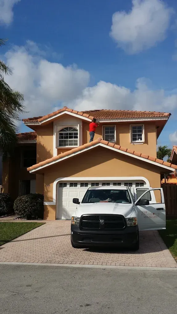 A person in a red shirt stands on the roof of a beige house, painting the trim near a white pickup truck in the driveway.