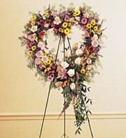 Floral heart-shaped wreath on a stand, with pink, yellow, and white flowers against a beige wall.