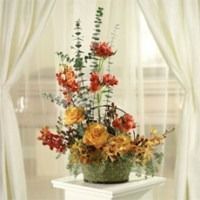 Tall floral arrangement with red, yellow, and orange flowers on a white table by a window